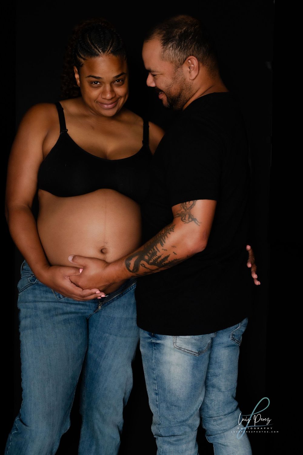 Tender maternity photo of a couple in jeans and black tops embracing against dark background.