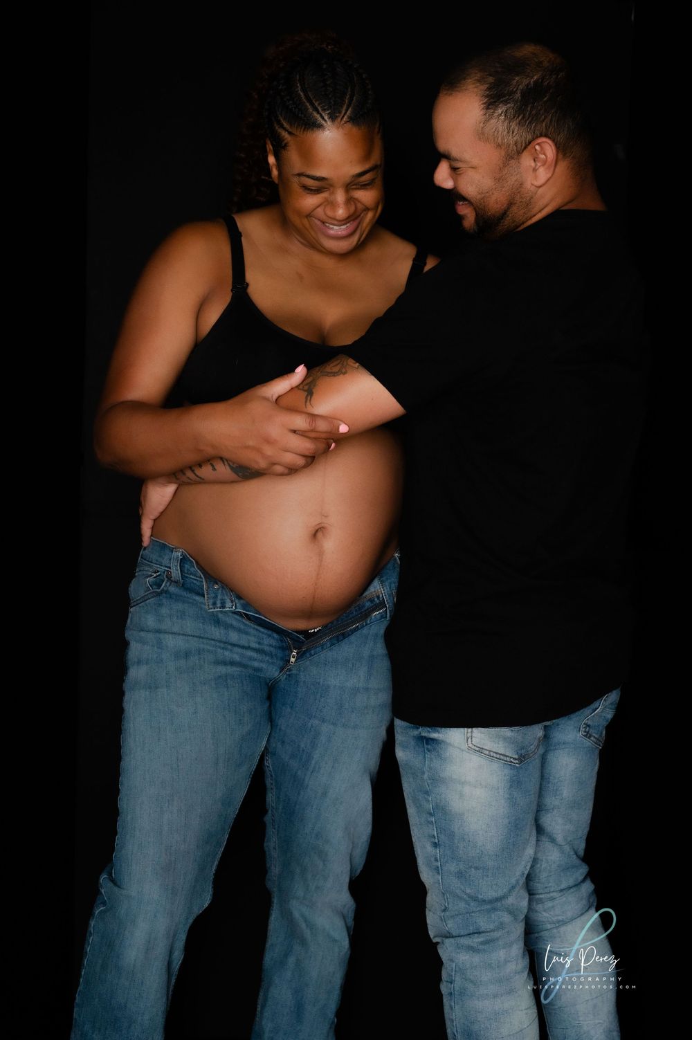 A couple wearing black and denim share a tender moment during a maternity photo shoot against a dark background.