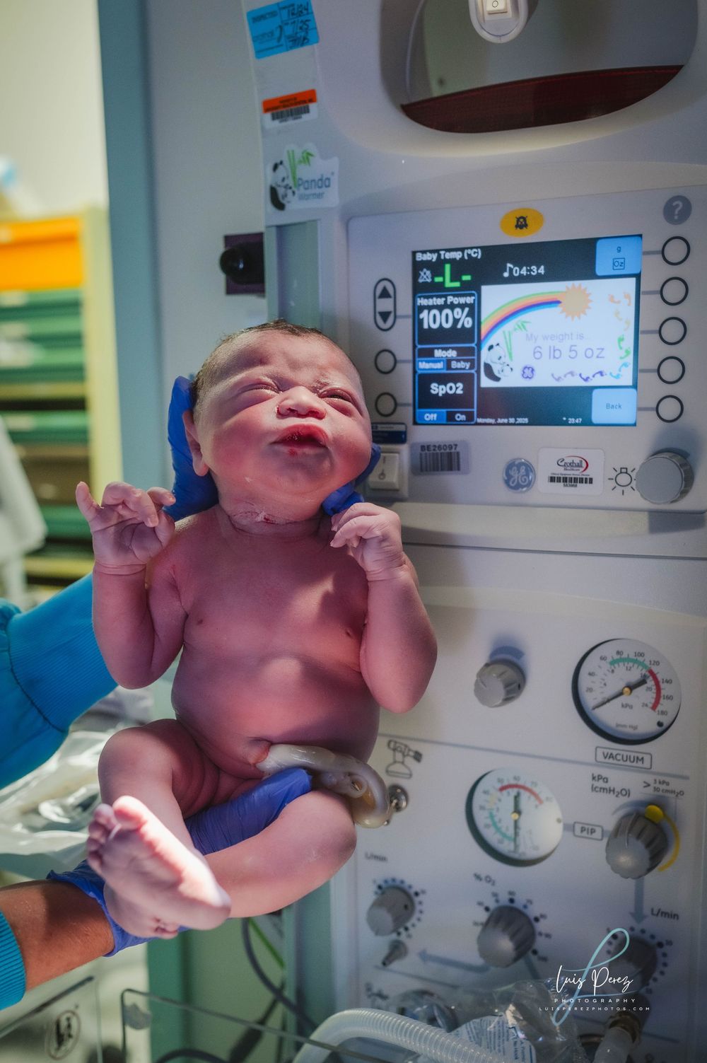Newborn baby in hospital delivery room with medical equipment and monitoring devices in background.