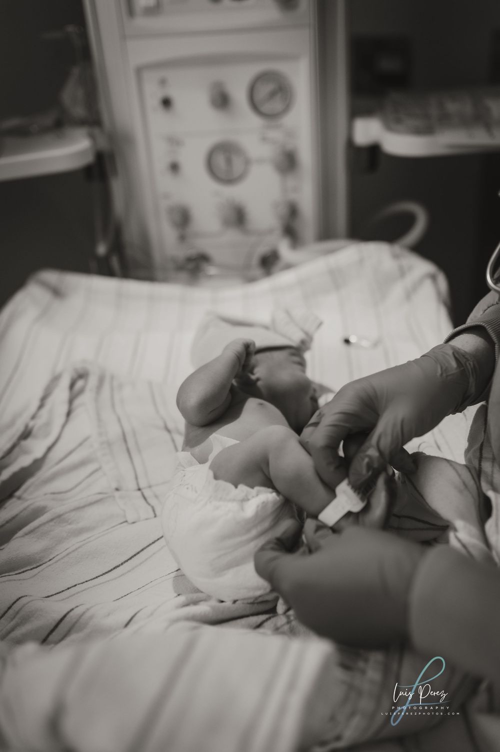 Black and white medical photo showing gloved hands examining a newborn baby on hospital bedding next to monitoring equipment.