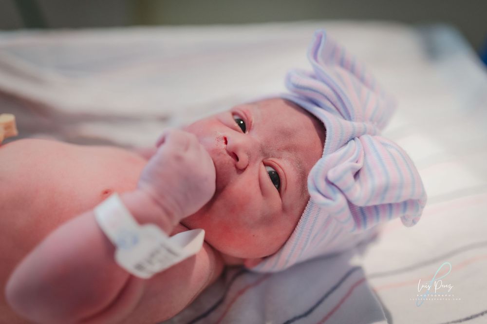 A newborn baby lying on white fabric wearing a light purple cap raises their tiny hand.