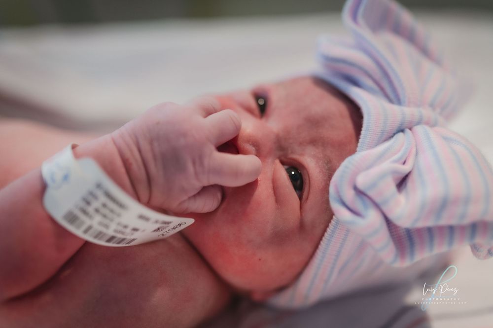 A newborn baby wearing a hospital ID bracelet and striped clothing is shown in close-up while sucking their thumb.