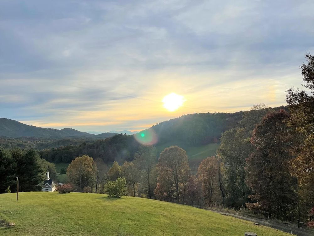 A scenic sunset over rolling hills with a white church steeple visible on a grassy slope surrounded by autumn trees.