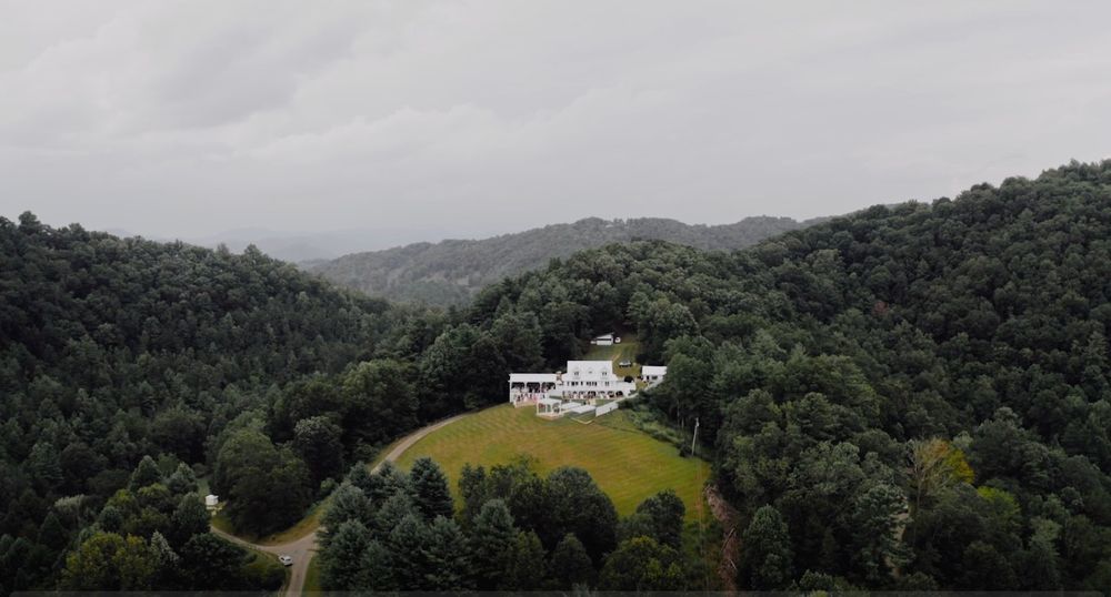 Aerial view of a modern white mansion perched on a hilltop surrounded by dense forest in a mountainous landscape.