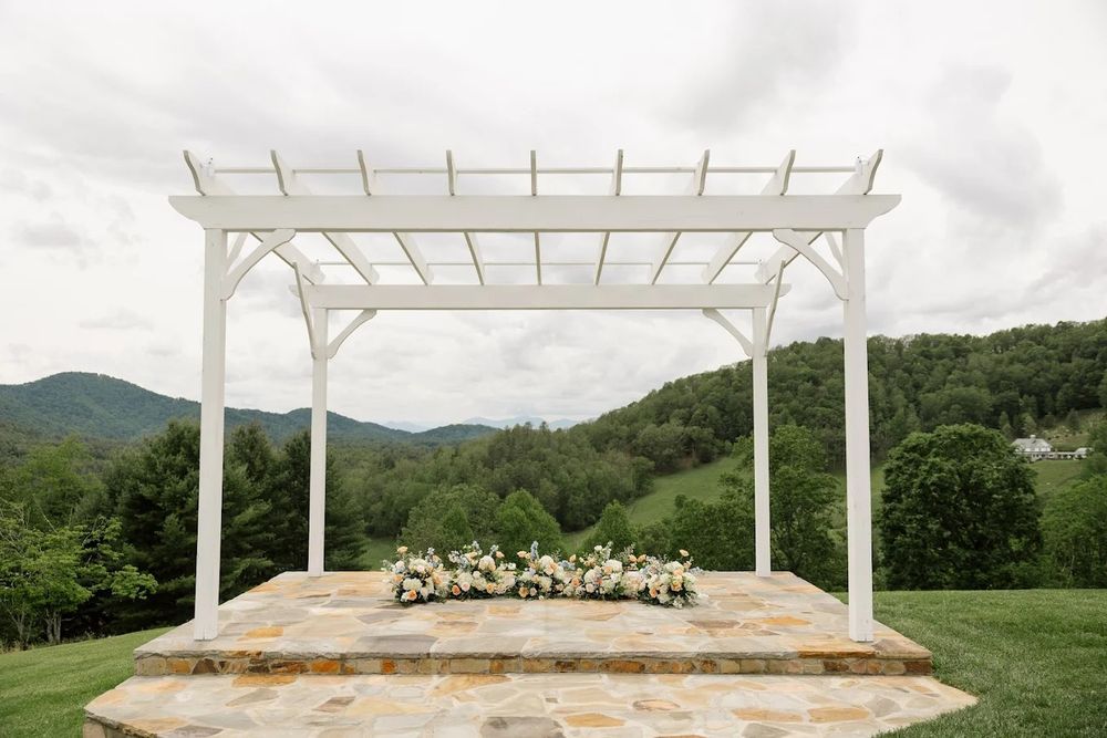 White wooden pergola structure set against a mountain landscape backdrop with lush green trees and cloudy sky.