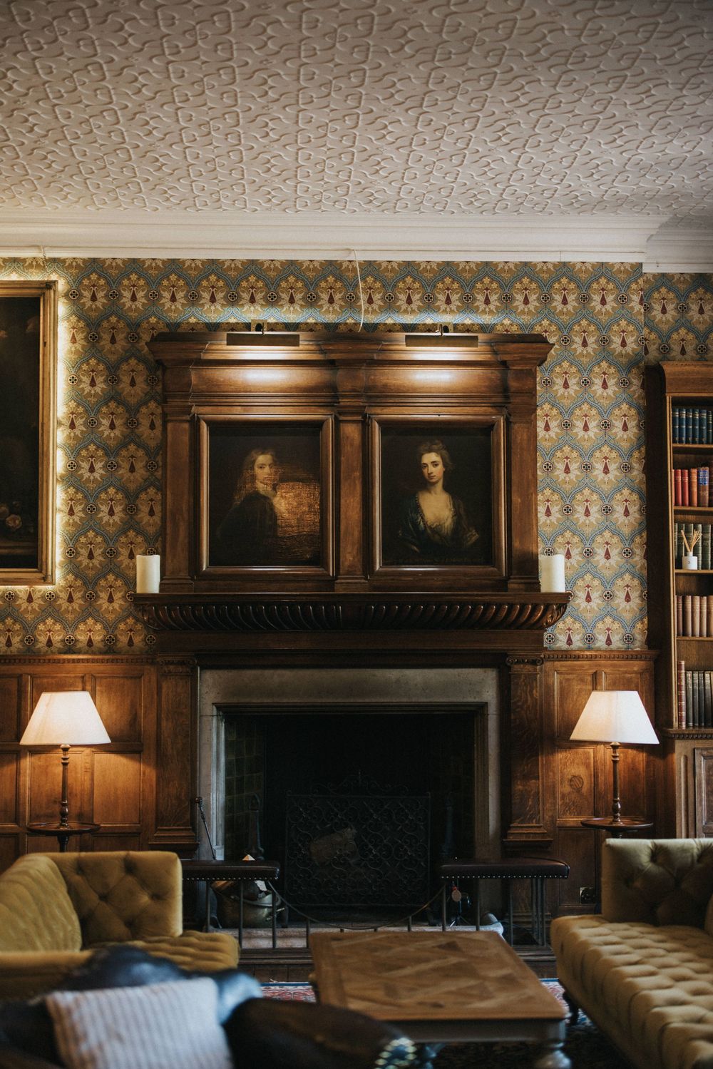 Elegant vintage library room with floral wallpaper, leather chesterfield sofas, antique portraits above fireplace and table lamps.