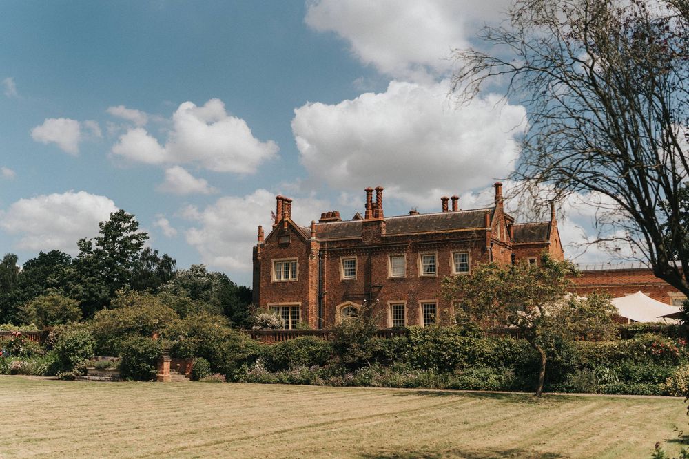 A grand Victorian red brick manor house with multiple chimneys stands behind a grassy lawn on a sunny day with white clouds.