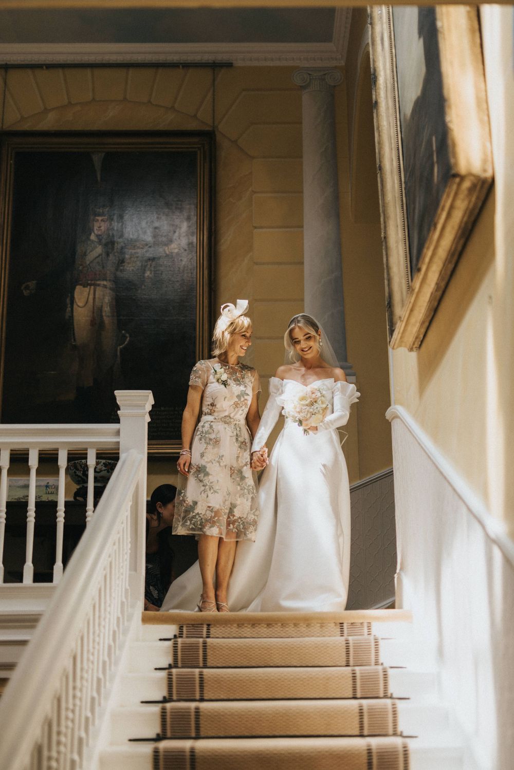 Two people in white dresses stand on elegant interior stairs in vintage-inspired architectural setting bathed in soft light.