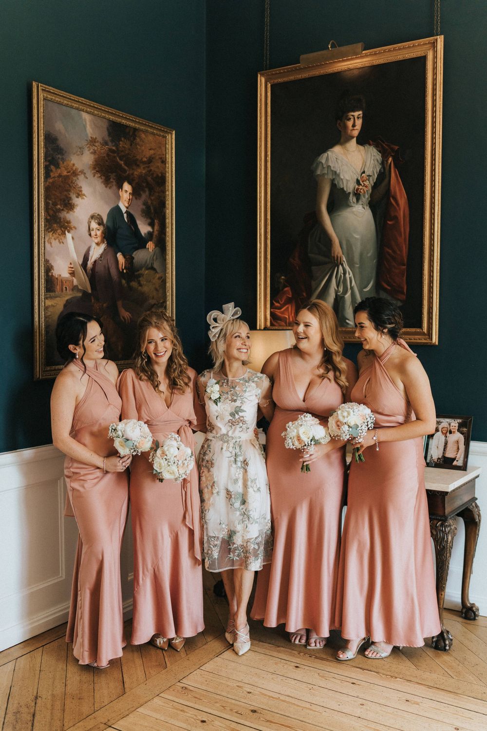 Bridesmaids in matching peach-colored dresses gather and laugh together in an elegant room with portrait paintings.