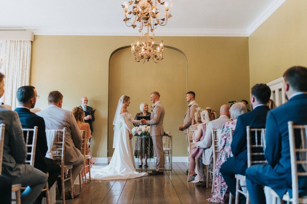Indoor wedding ceremony in an elegant room with arched doorway and chandelier showing a couple and officiant at the altar.