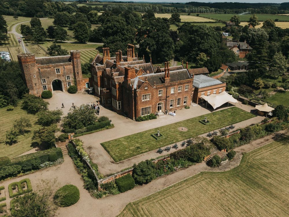 Aerial view of a grand red brick manor house with ornate architecture surrounded by manicured gardens and countryside.