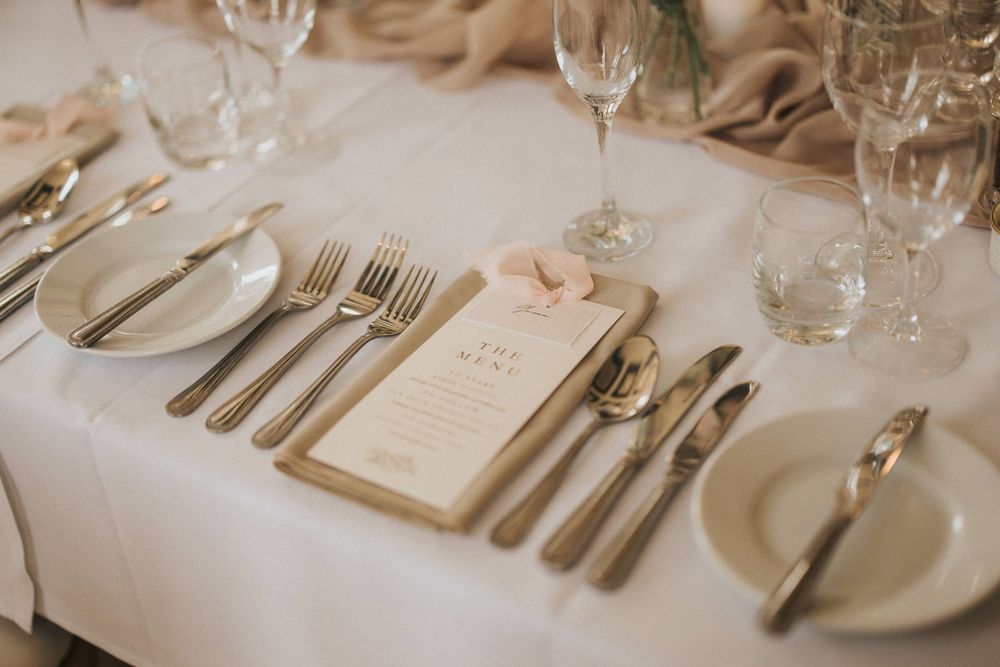 Elegant formal place setting with white linens, silverware, menu card, and wine glasses at a wedding reception.