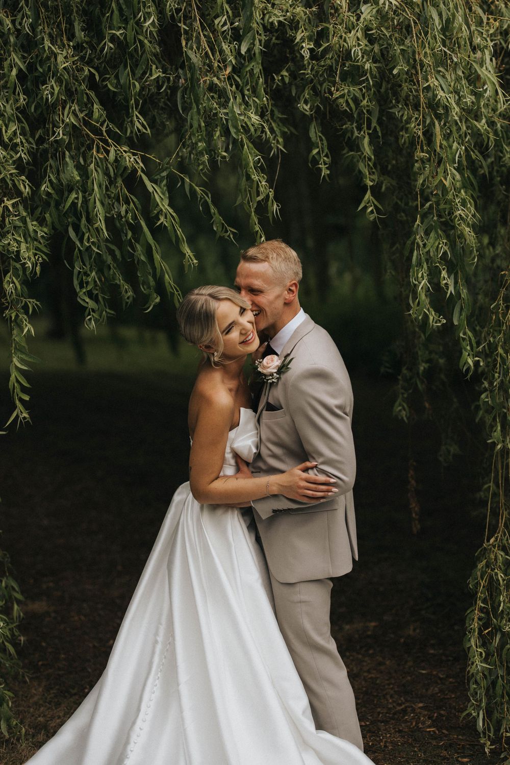 A romantic moment captured between two people in wedding attire sharing kisses under a weeping willow tree.