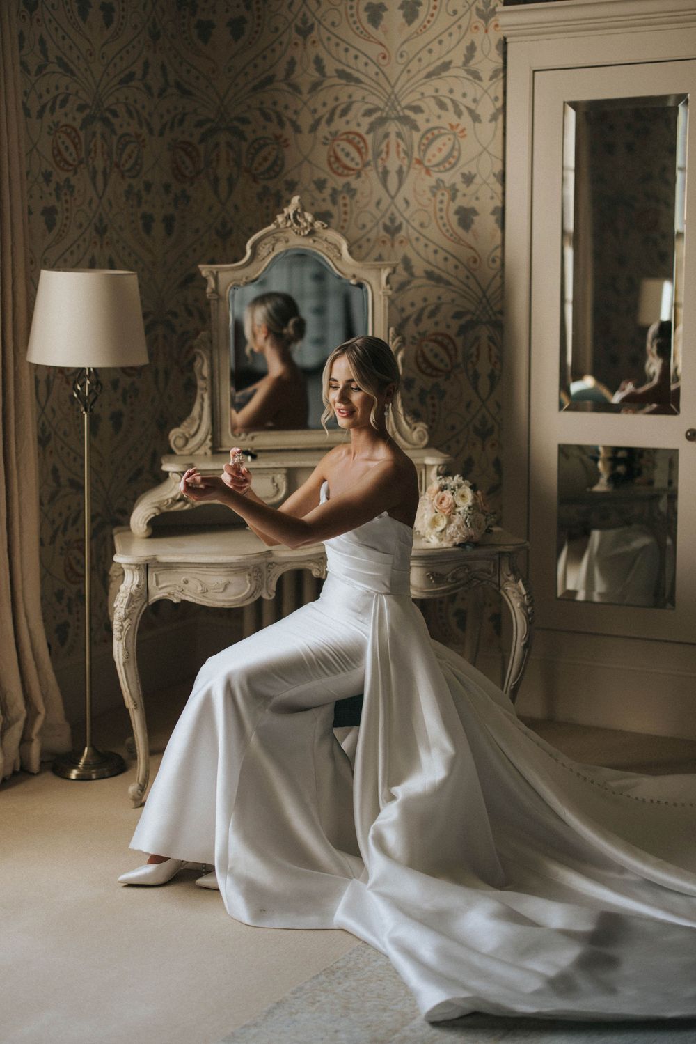 Elegant bride in flowing white gown sits at ornate vintage vanity with floral wallpaper and lamp in reflection.