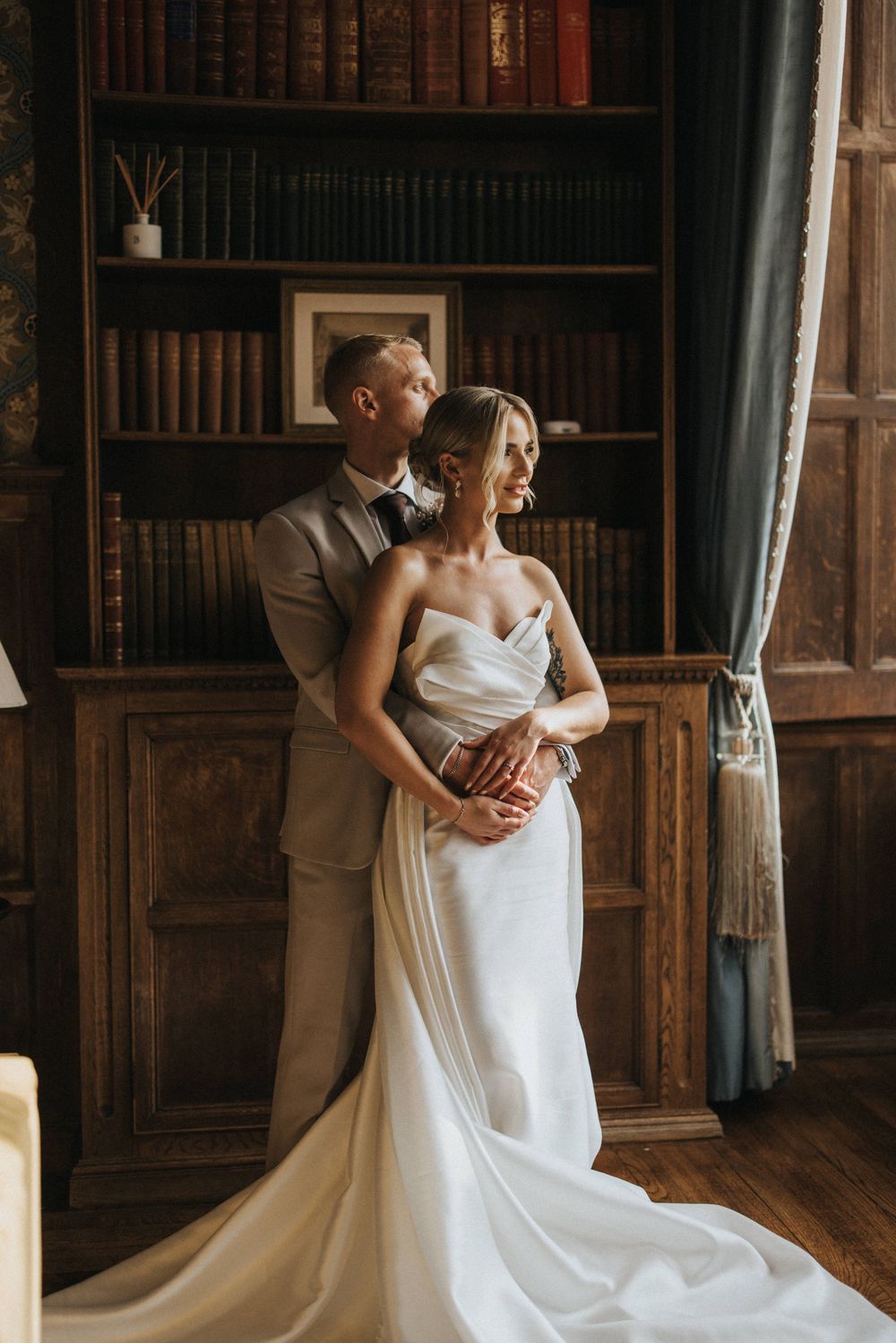 Newlywed couple embraces in romantic moment inside vintage library with wooden bookshelves.