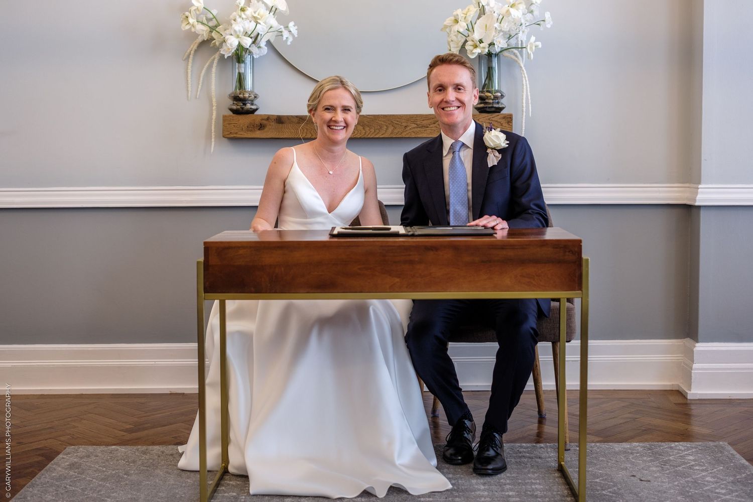 A happy couple sits together at an elegant wooden desk with white floral arrangements during their wedding ceremony.