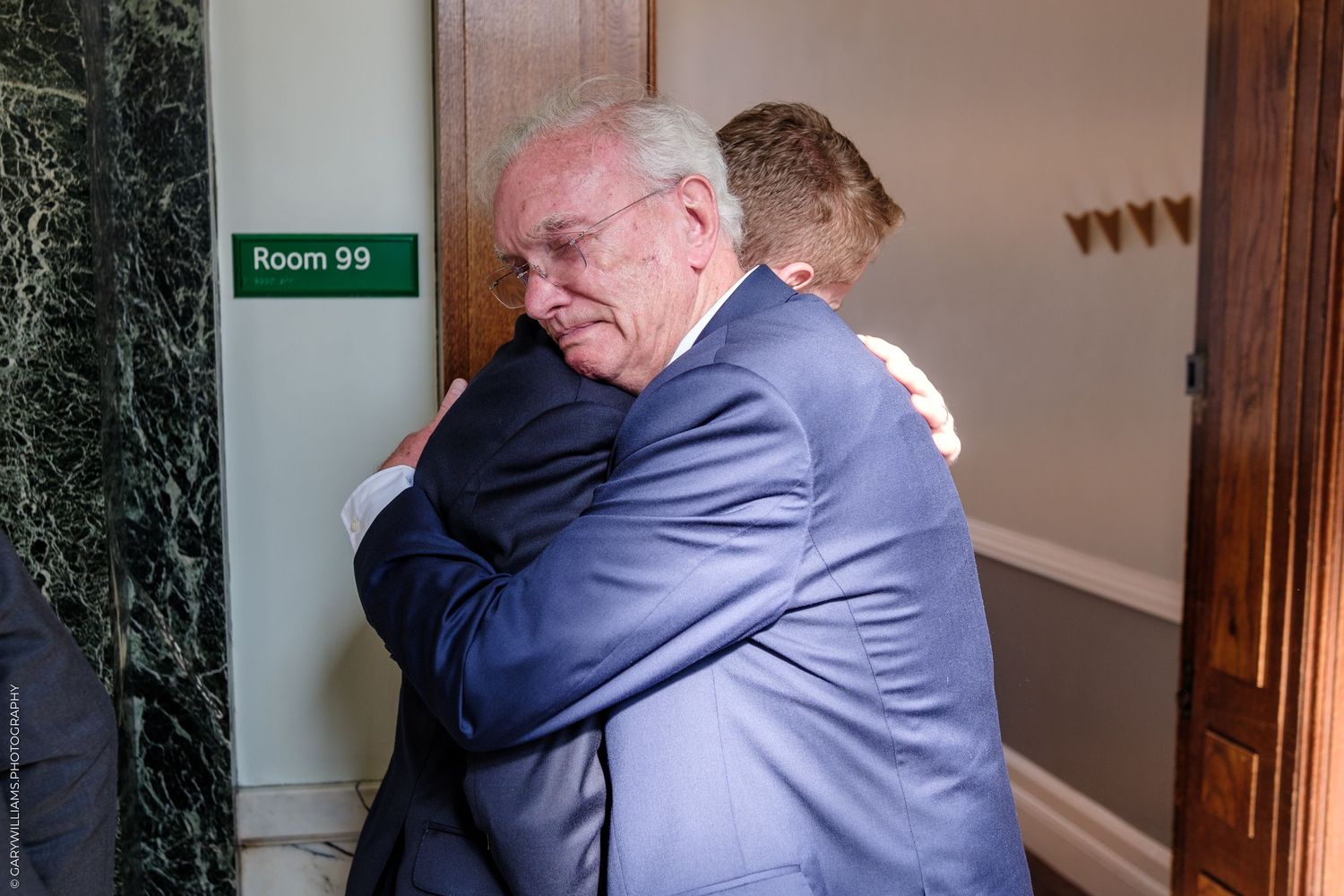 Two people share an emotional embrace in a hallway near Room 99.
