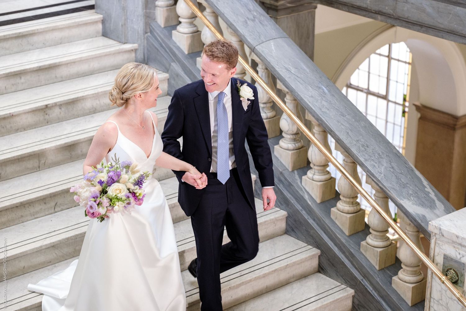 A couple descends an elegant marble staircase together, with the bride holding a vibrant bouquet.