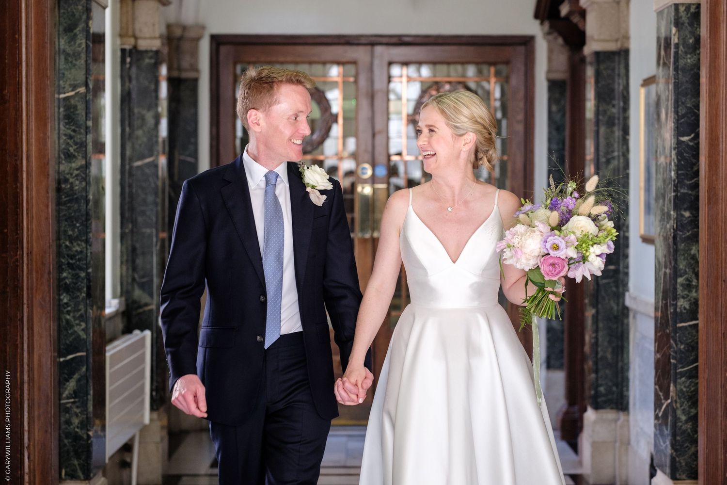 Happy newlywed couple holding hands and laughing while walking through an elegant marble hallway after their wedding ceremony.