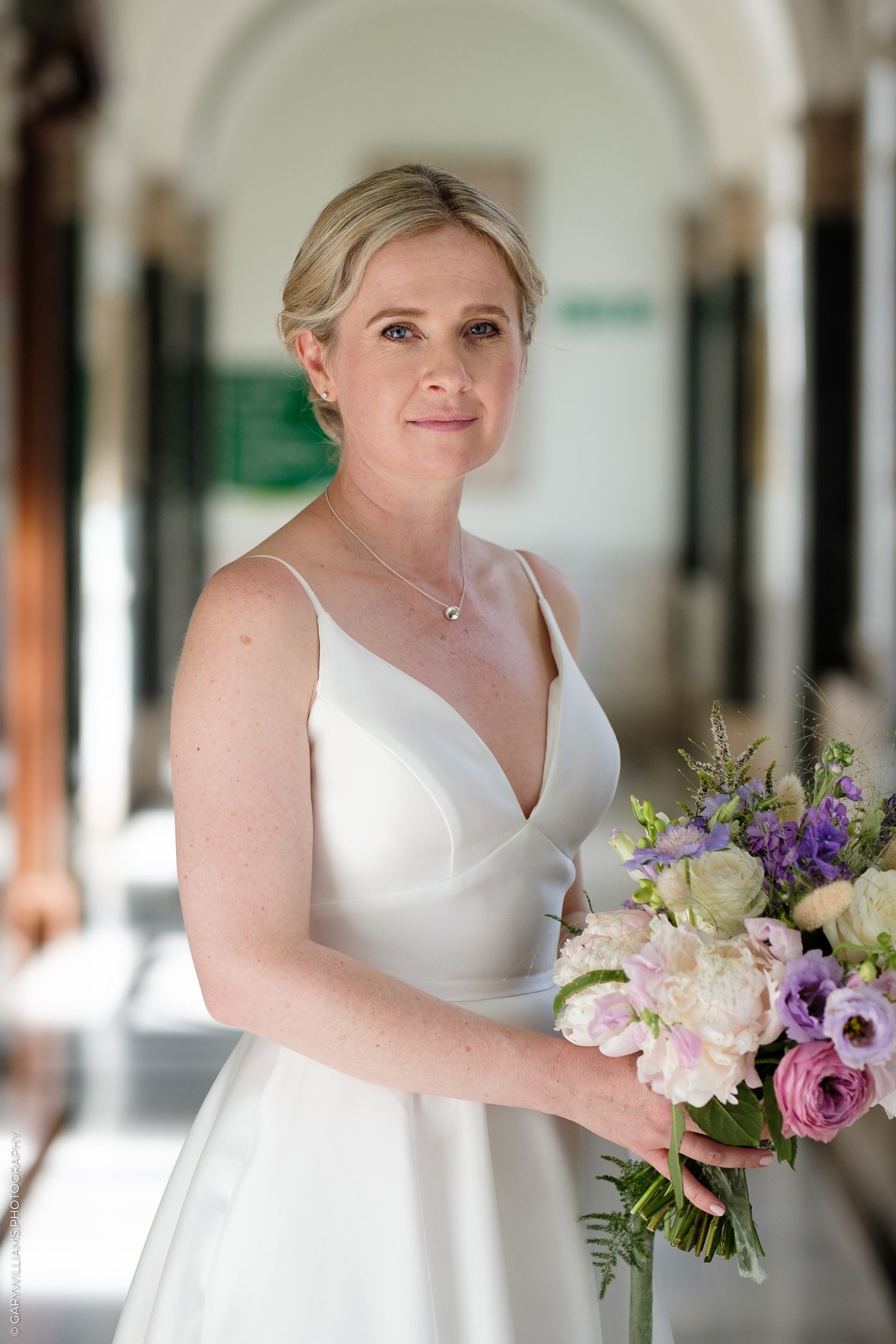 A bride in white dress holds a colorful bouquet of pink and purple flowers in an elegant hallway with arched ceilings.
