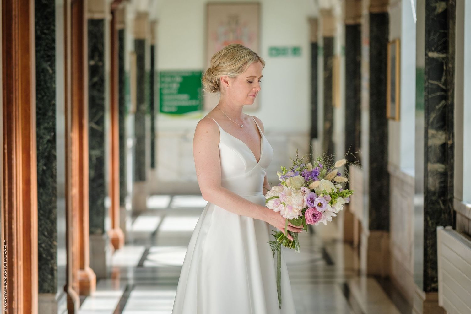 A bride in an elegant white sleeveless gown holds a pastel bouquet while standing in a bright hallway.
