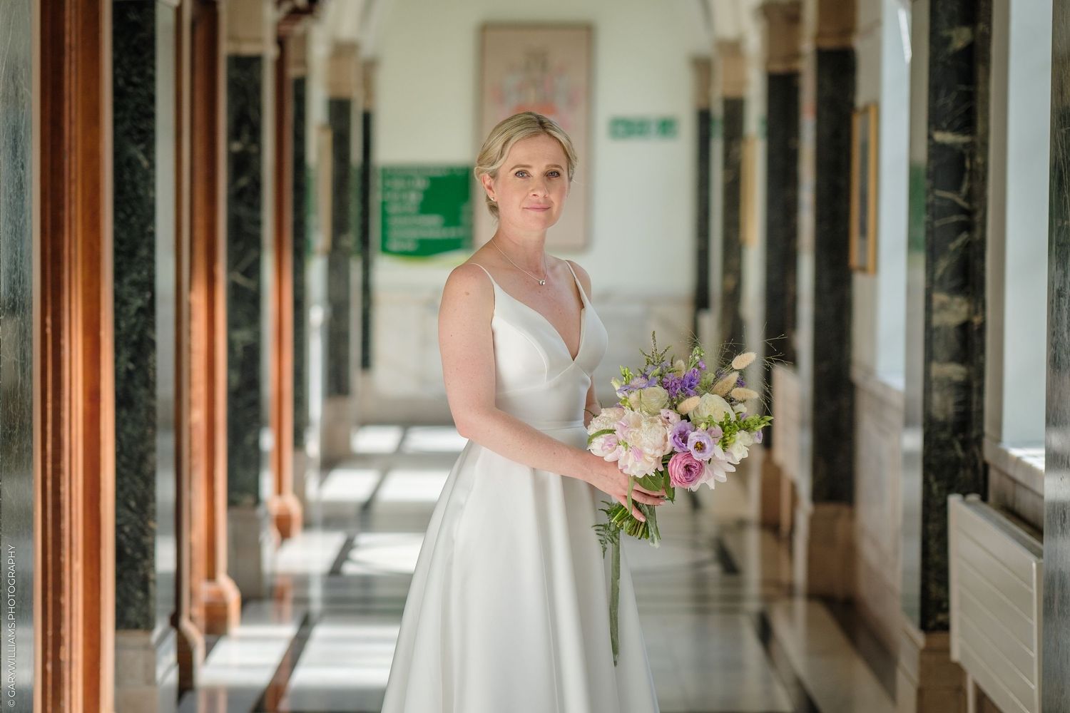A person wearing a white wedding dress walks through an elegant hallway with wooden pillars and natural lighting.