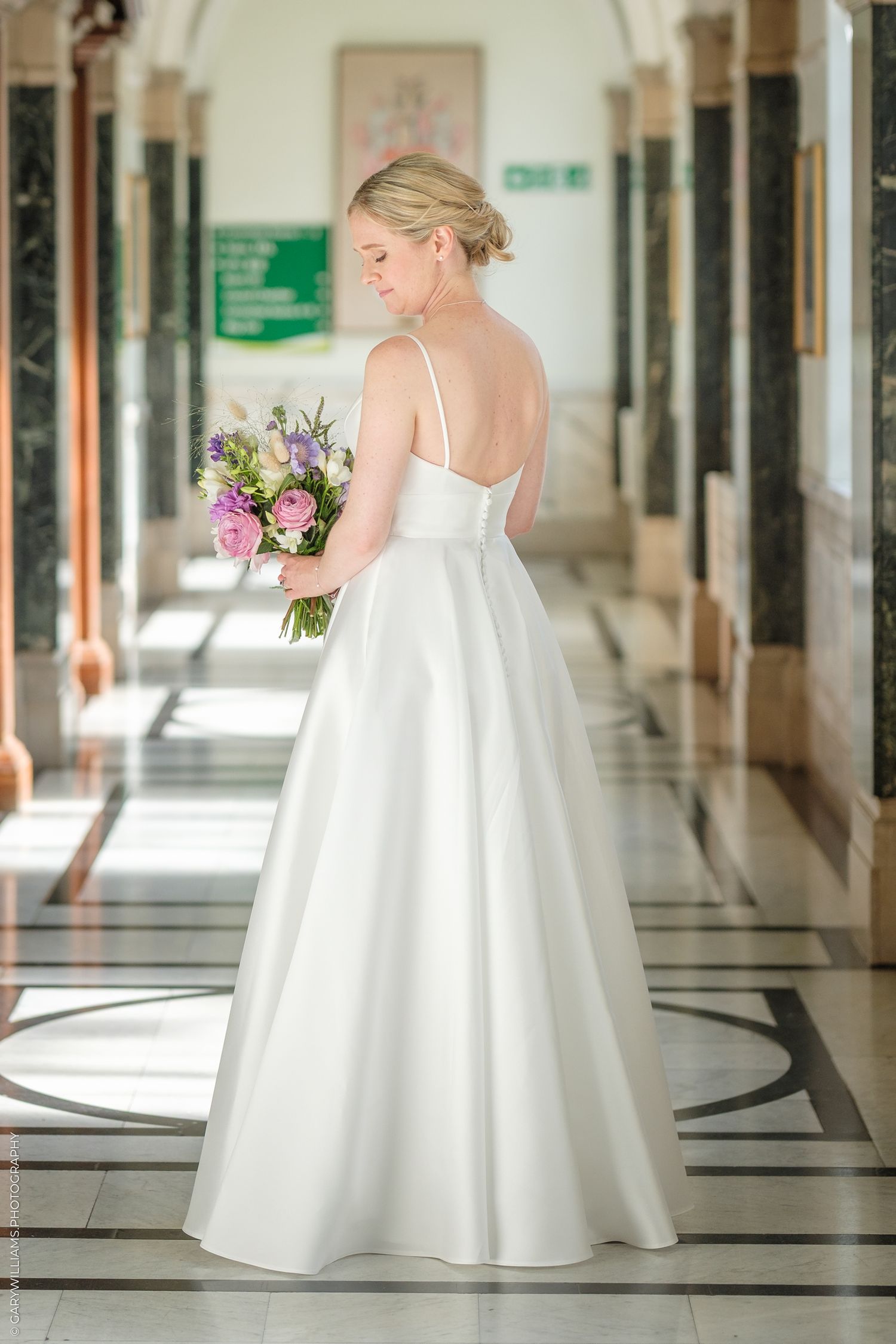 Elegant bride in simple white satin wedding gown holds colorful bouquet while standing in ornate hallway.