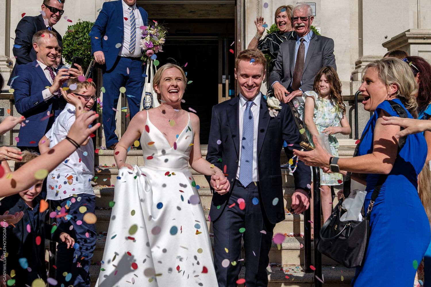 Wedding guests shower newlyweds with colorful confetti on the stone steps of a building during daytime celebration.