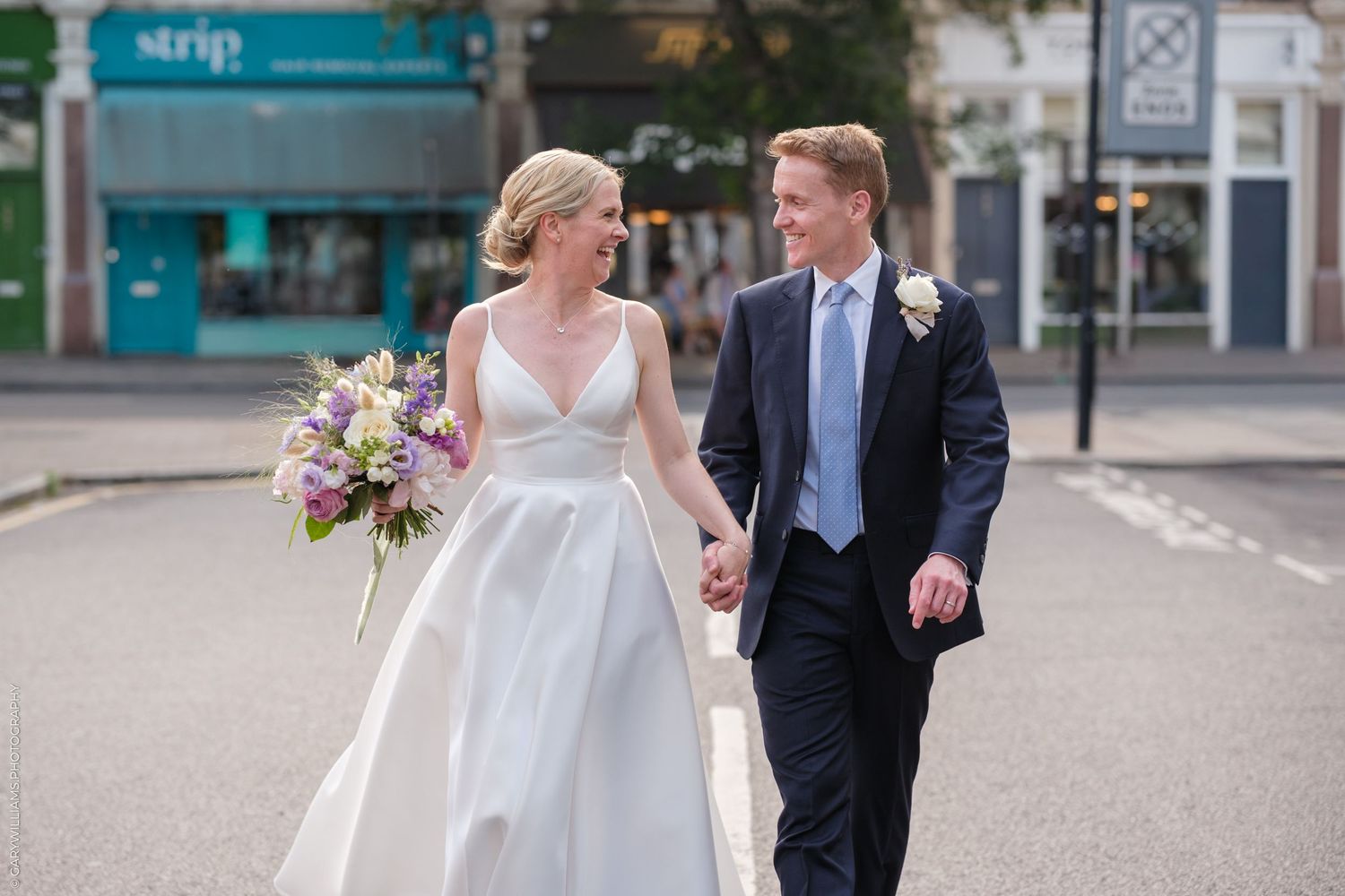 Bride in flowing white gown carries purple bouquet while walking with groom in navy suit on city street.