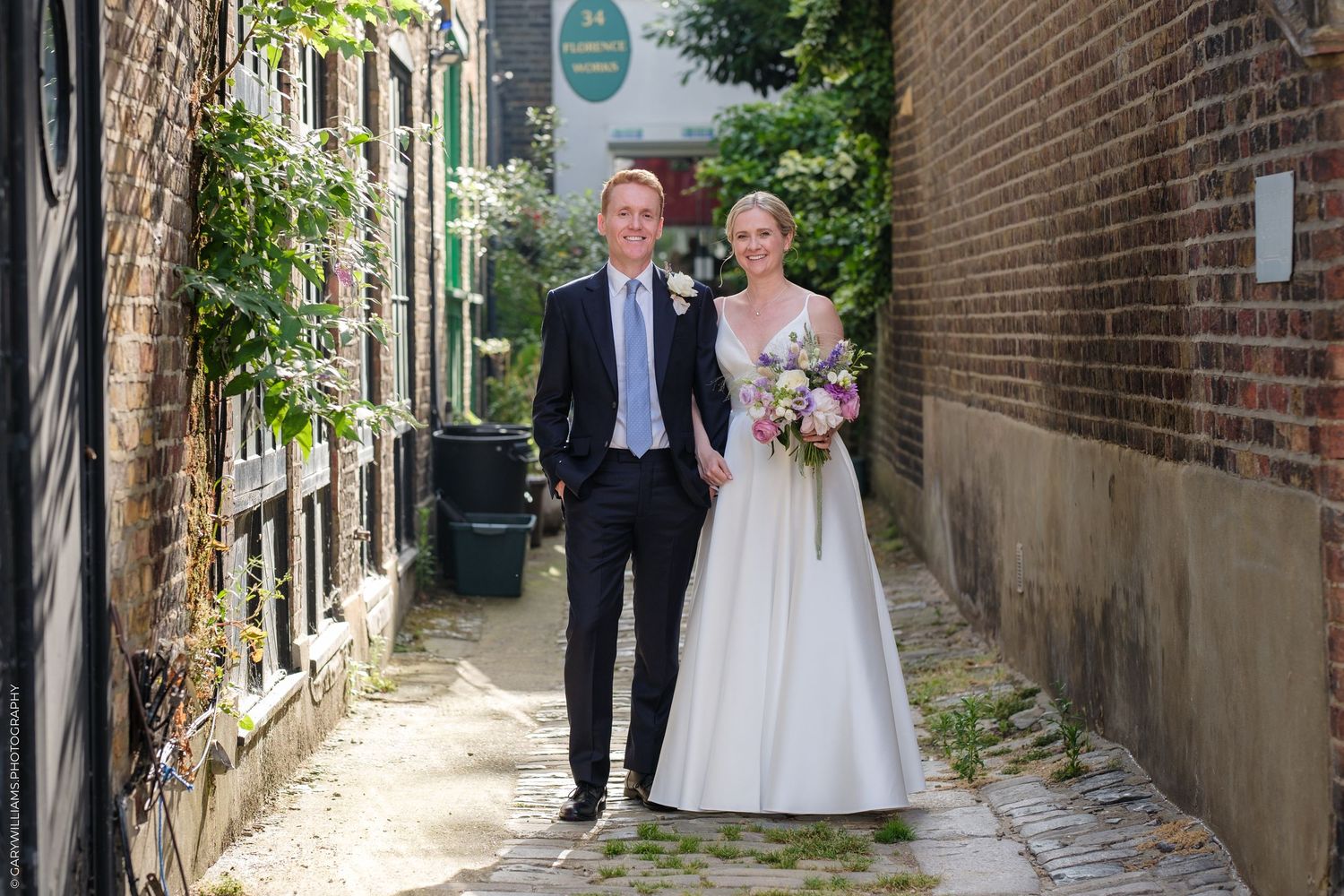A couple poses together in formalwear on a charming cobblestone alley between brick buildings on their wedding day.