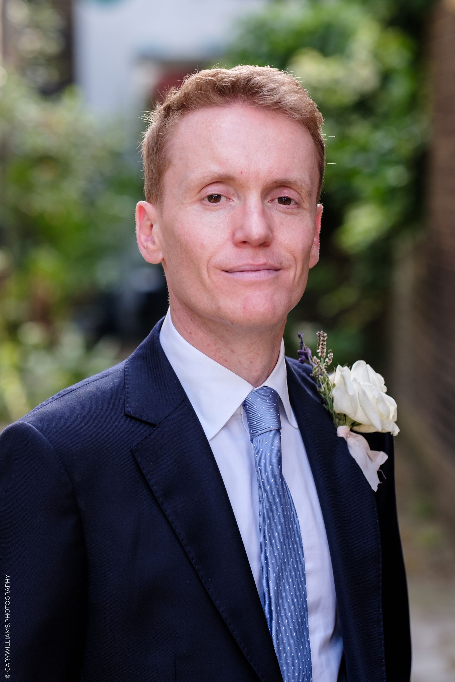 A man in a formal black suit and blue tie wearing a white boutonniere smiles at the camera.
