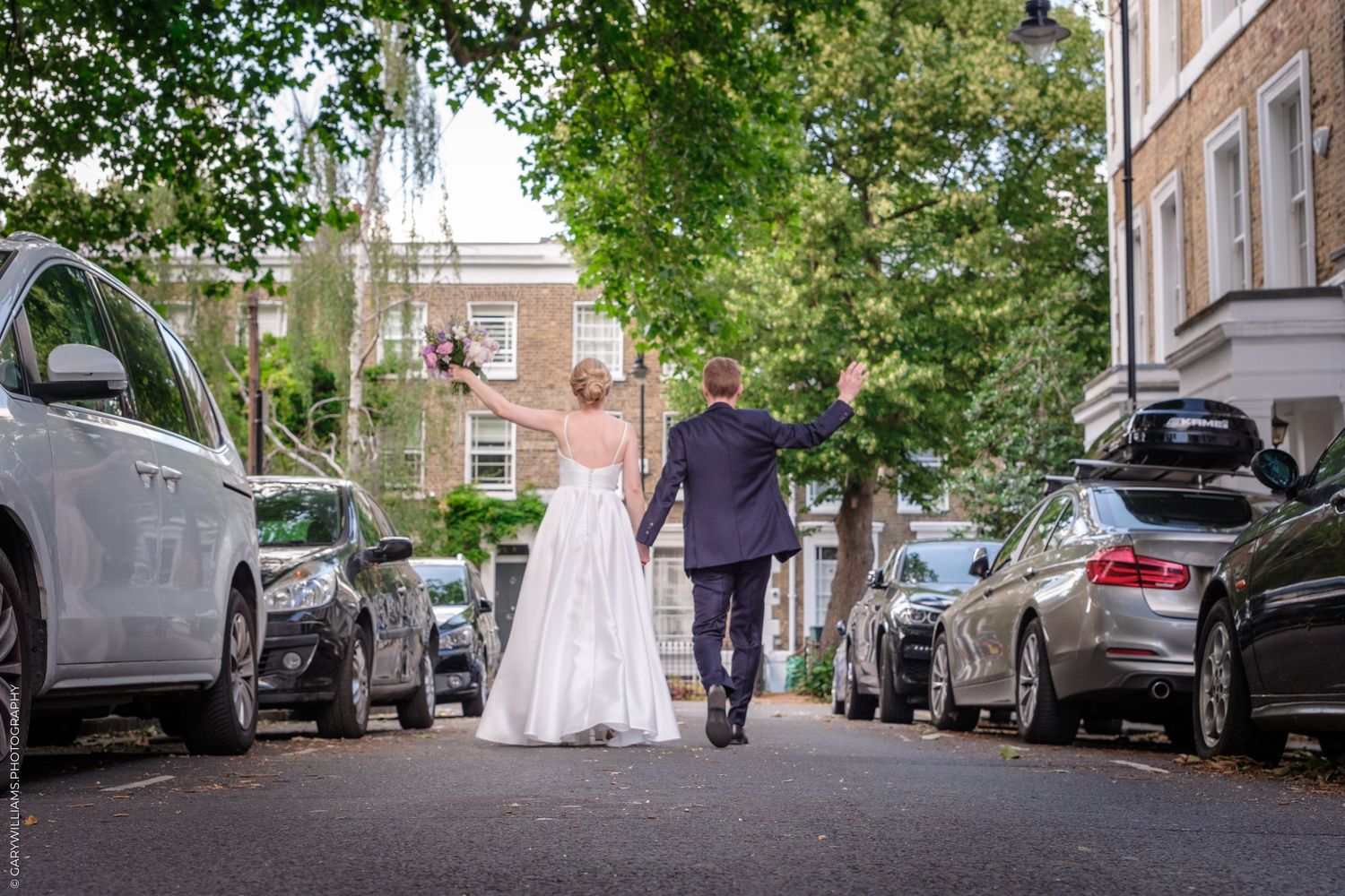 A couple in wedding attire walks down a tree-lined street between parked cars on their special day.