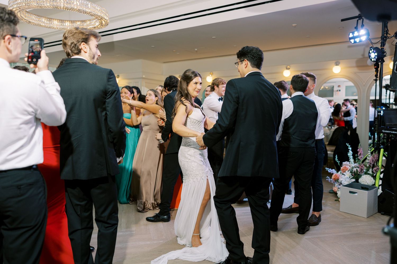 People dancing and socializing at an elegant formal event in a ballroom with a couple in white attire at center.