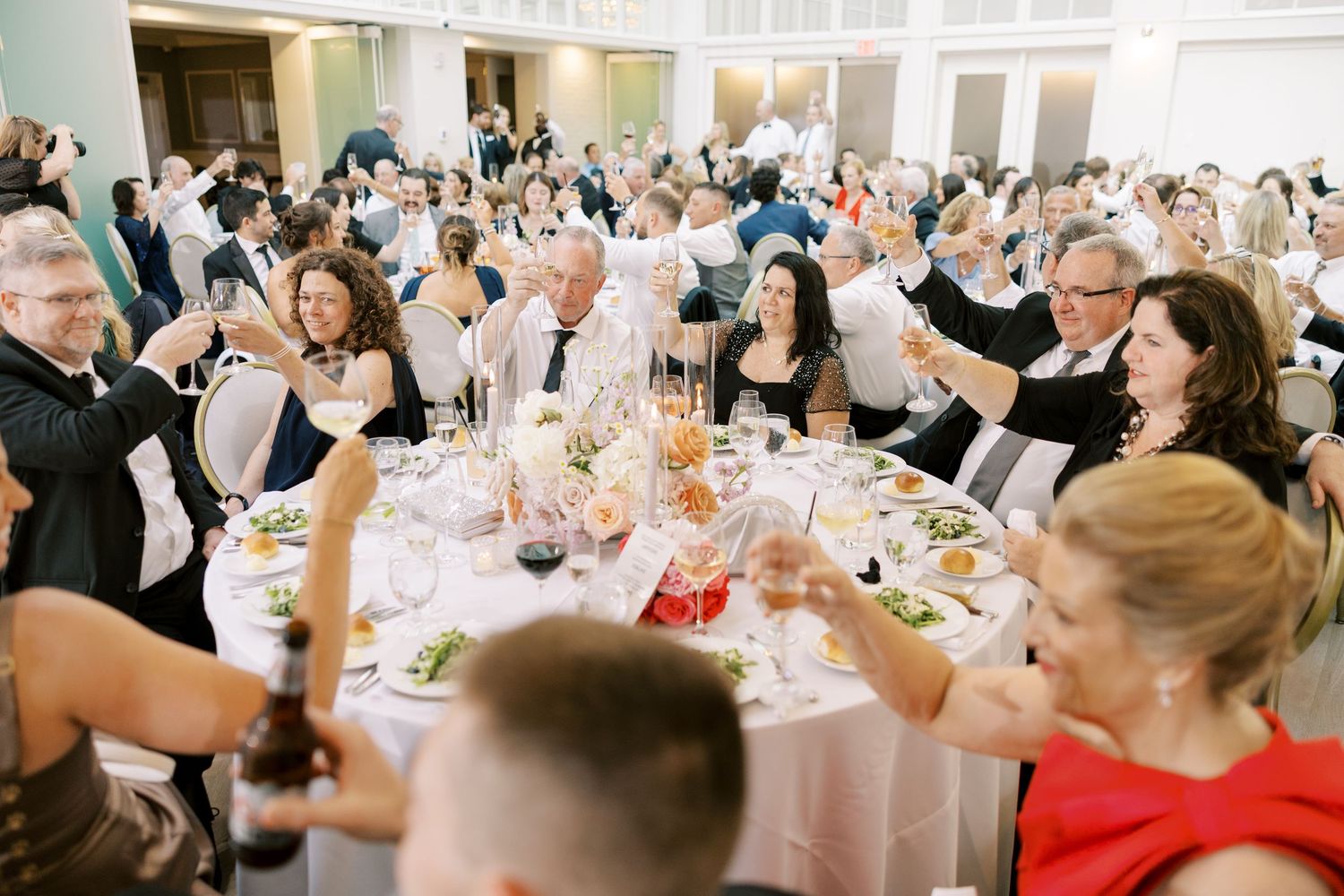 Wedding guests raise their glasses in a toast during an elegant reception with round tables and light pink decor.