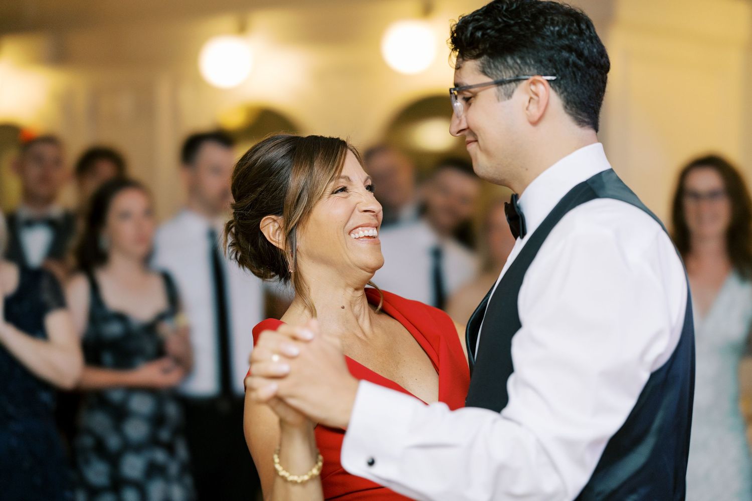 A couple in formal attire dance together at an event while guests look on from behind.