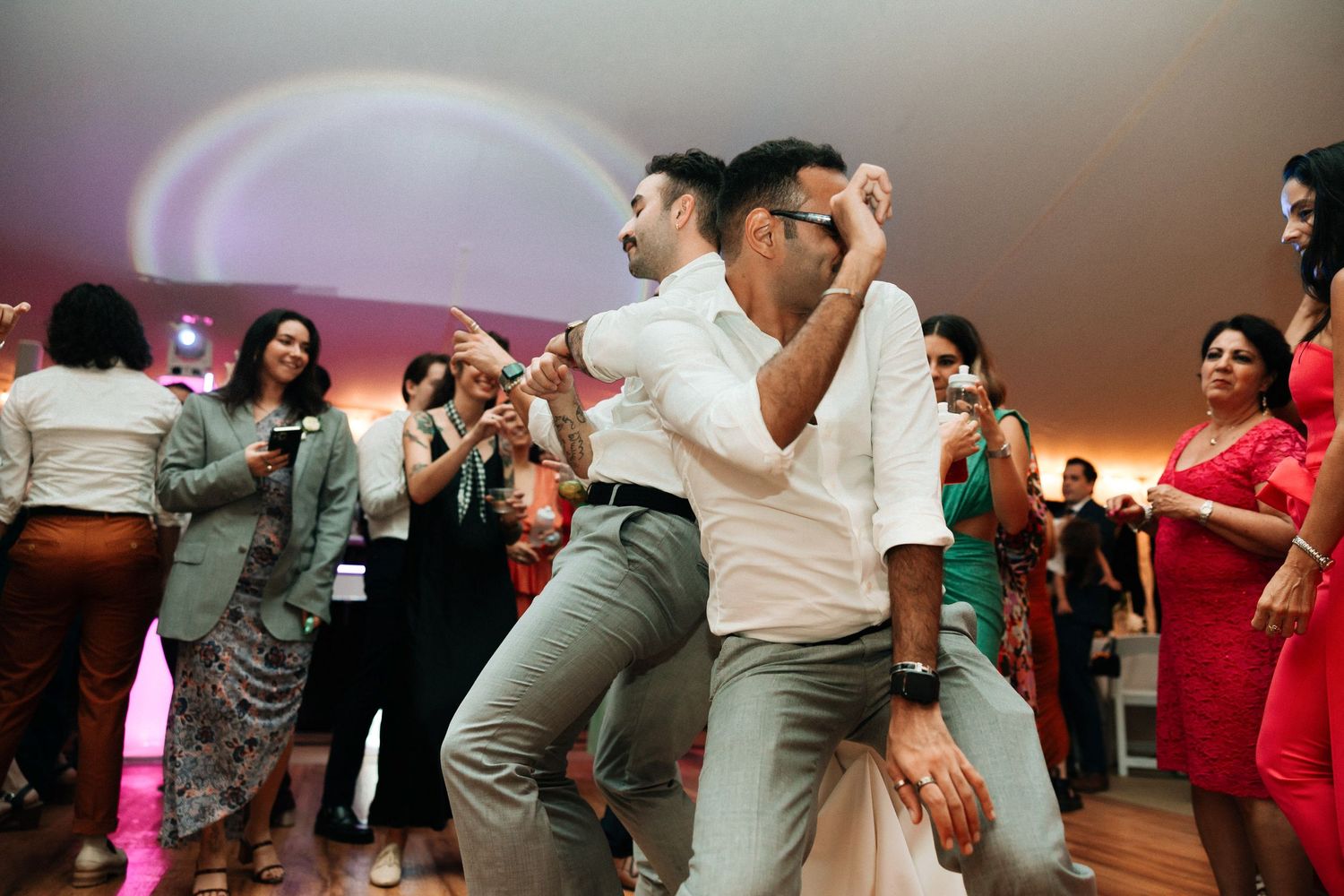 Wedding guests dance enthusiastically on a crowded reception dance floor with colorful mood lighting in the background.