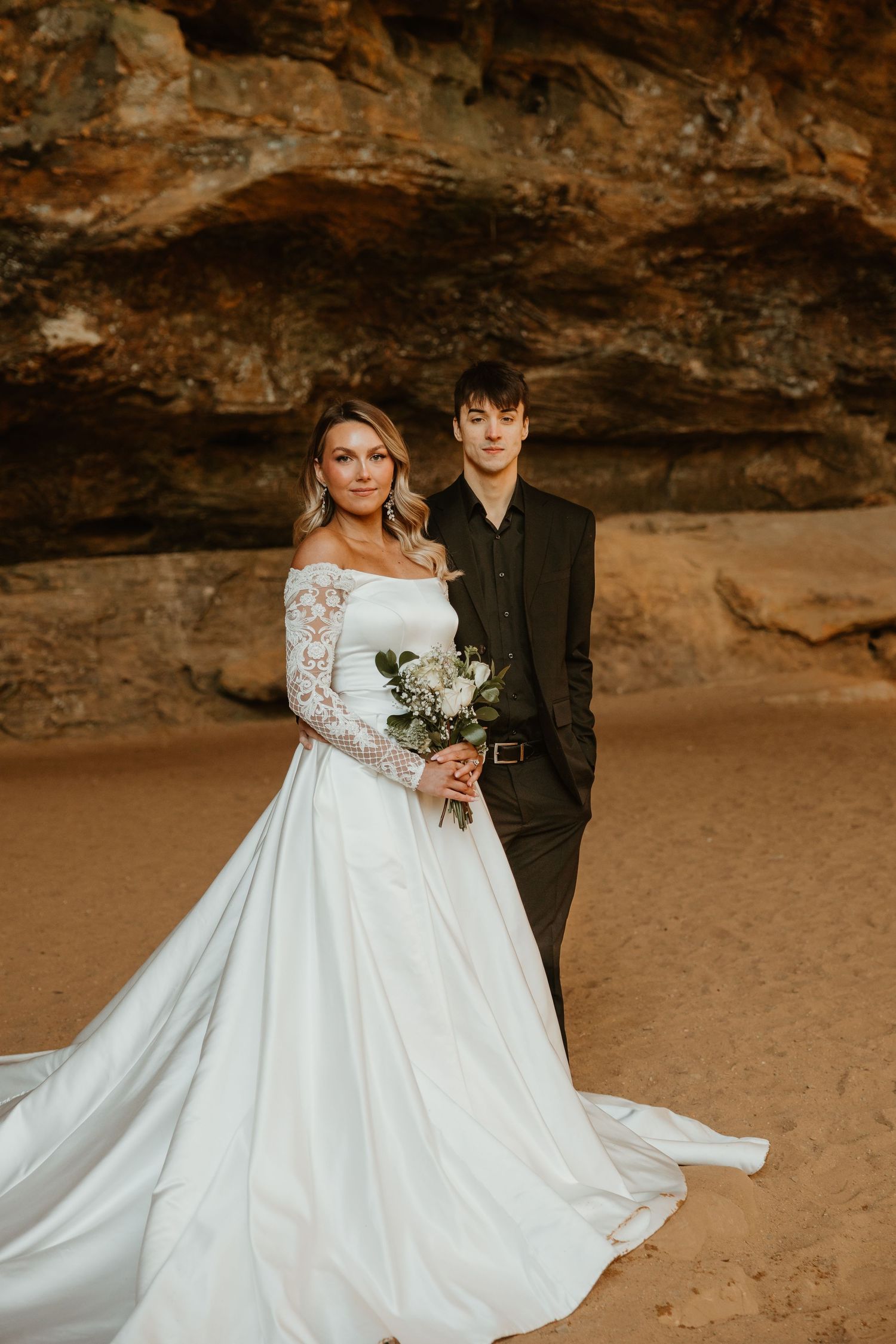 Wedding couple poses in off-white gown and black suit against dramatic rock cave formation in golden light.