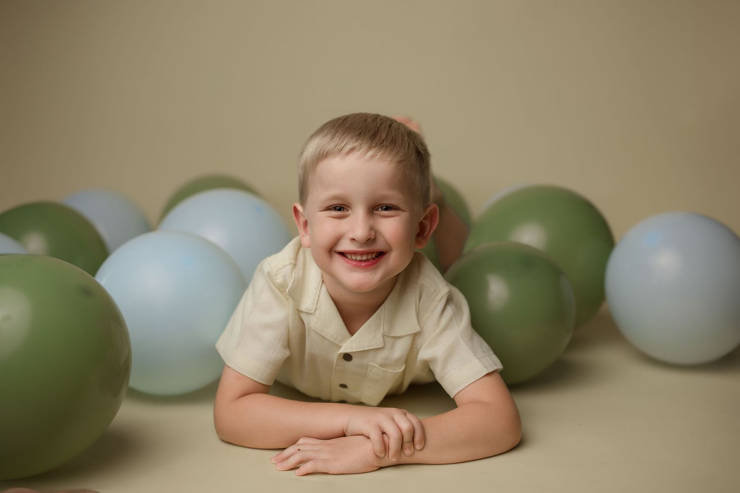 Young child laying on floor smiling with soft green and blue balloons in background during photo session.