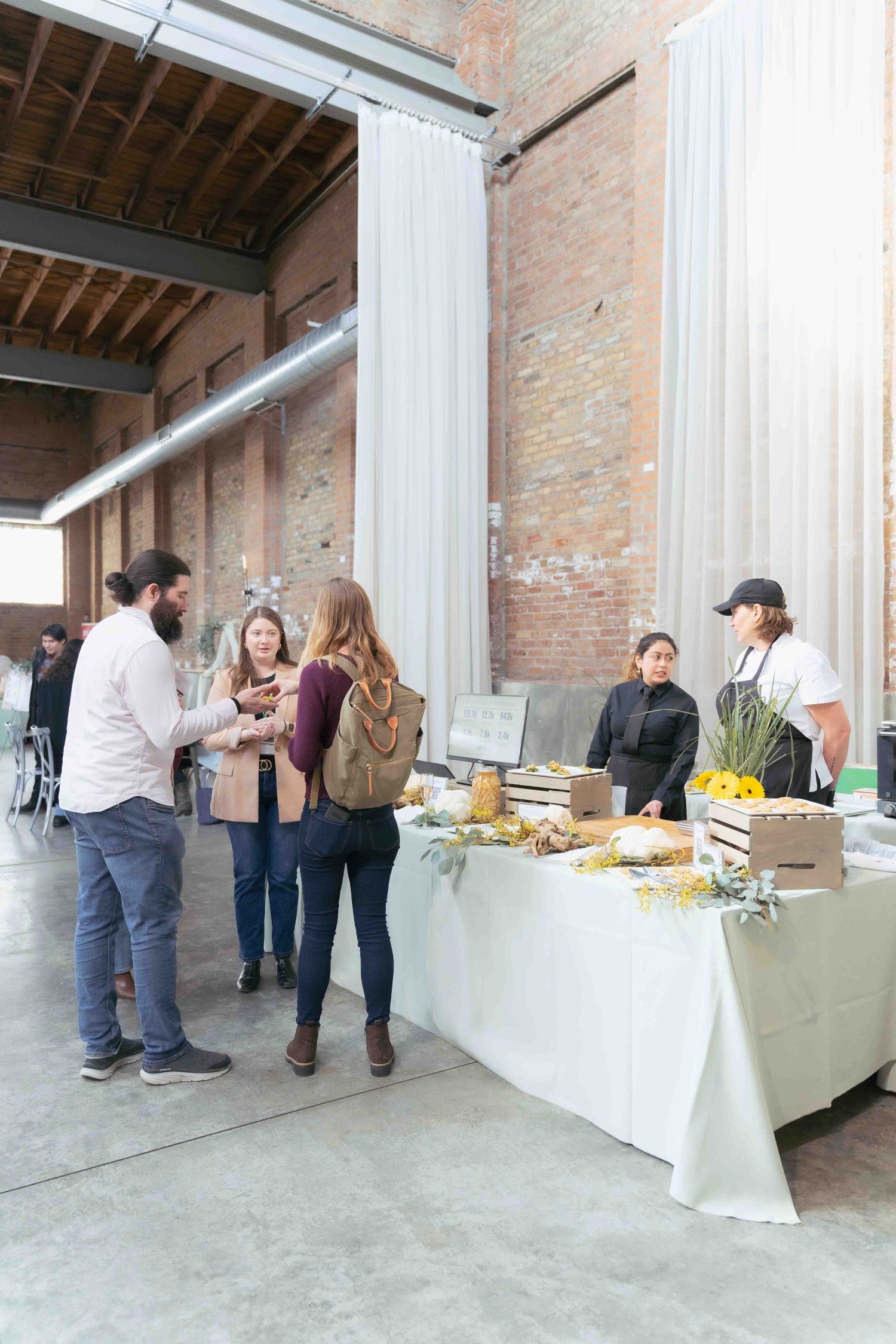 Group of attendees gathering around a catering table at an industrial-style venue with exposed brick walls. At Fairlie, Chicago
