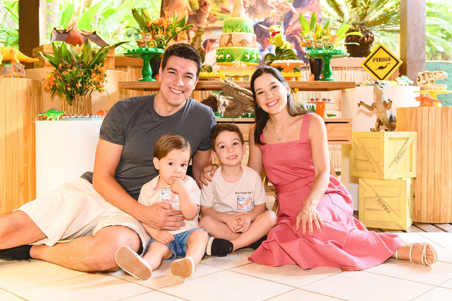 Family sits together on floor of bright sunlit room surrounded by tropical plants and wooden home decor.
