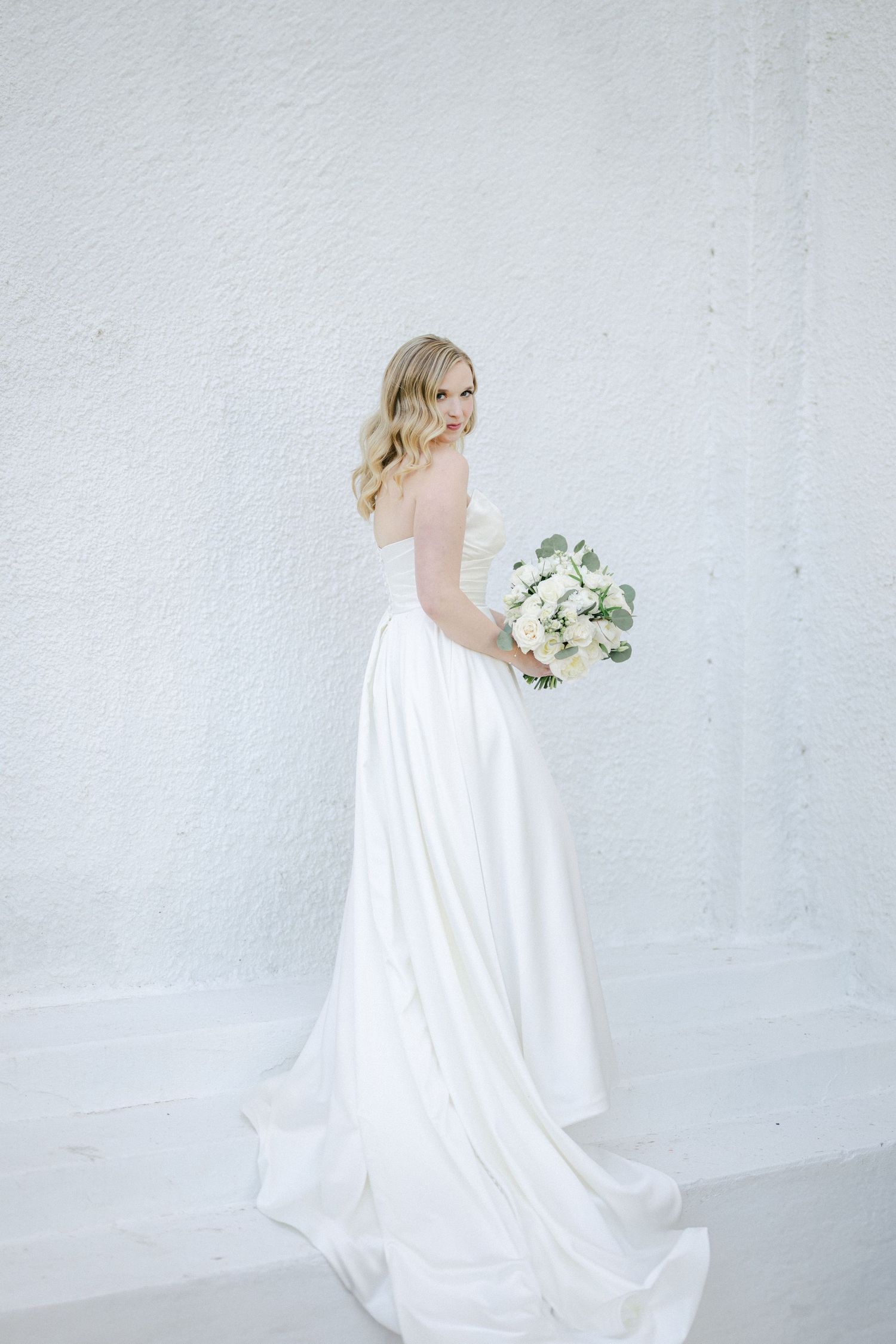 Elegant bride in flowing white gown holds a soft white and green floral bouquet against minimalist white backdrop.