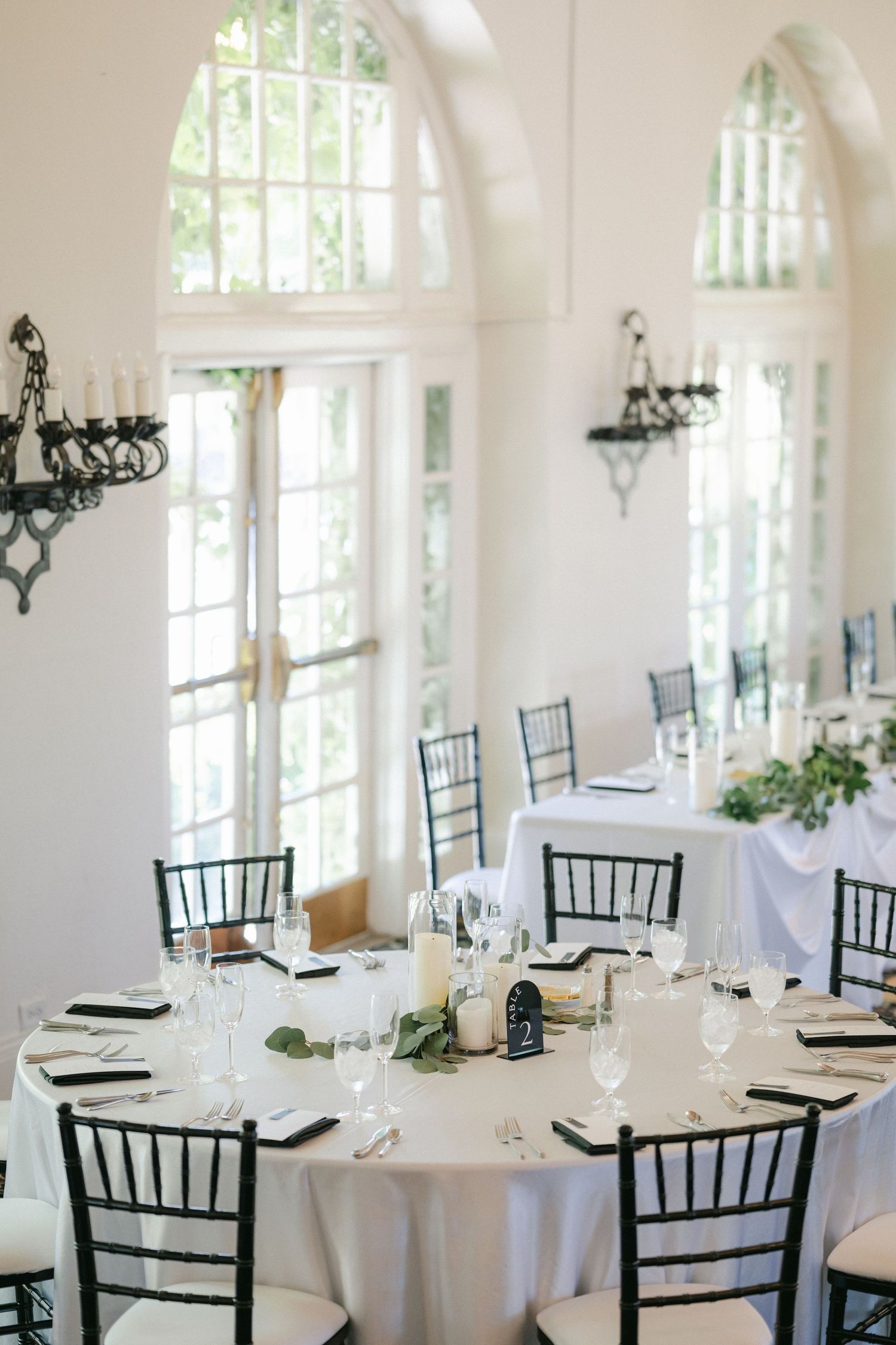 White tablecloths cover round tables in an elegant ballroom with arched windows and ornate chandeliers.