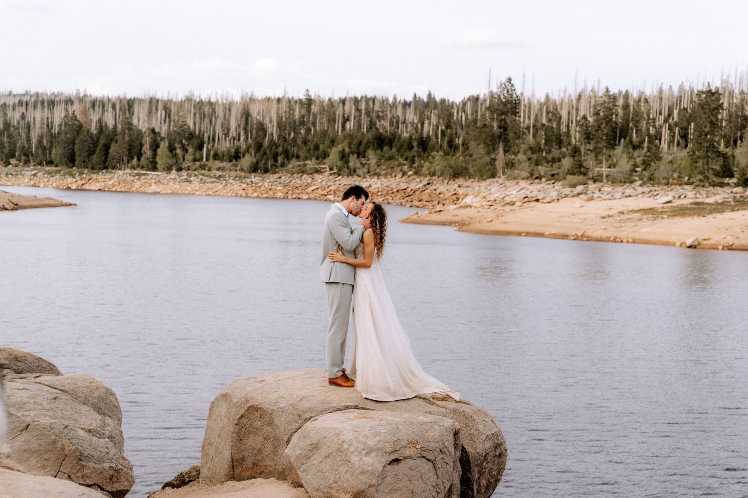 A couple embraces on large rocks by a serene lake with forest in the background.
