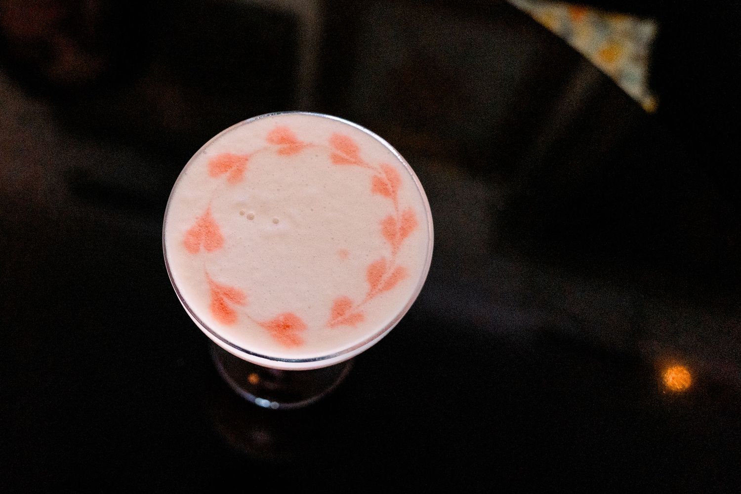 A creamy cocktail in a coupe glass with delicate pink heart-shaped designs floating on the foam surface.