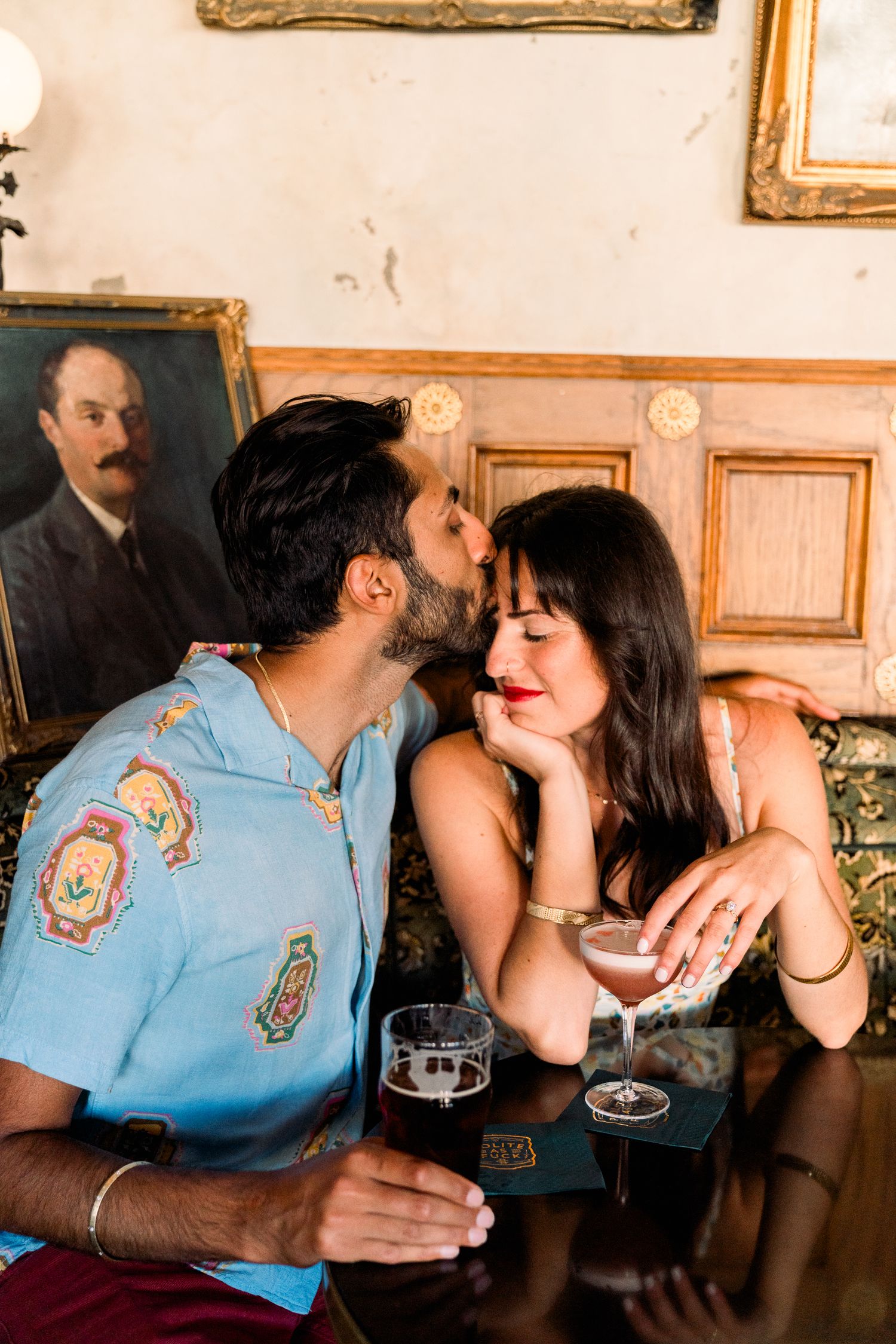 Romantic couple sharing intimate moment at bar with drinks and vintage portrait on wall in background.