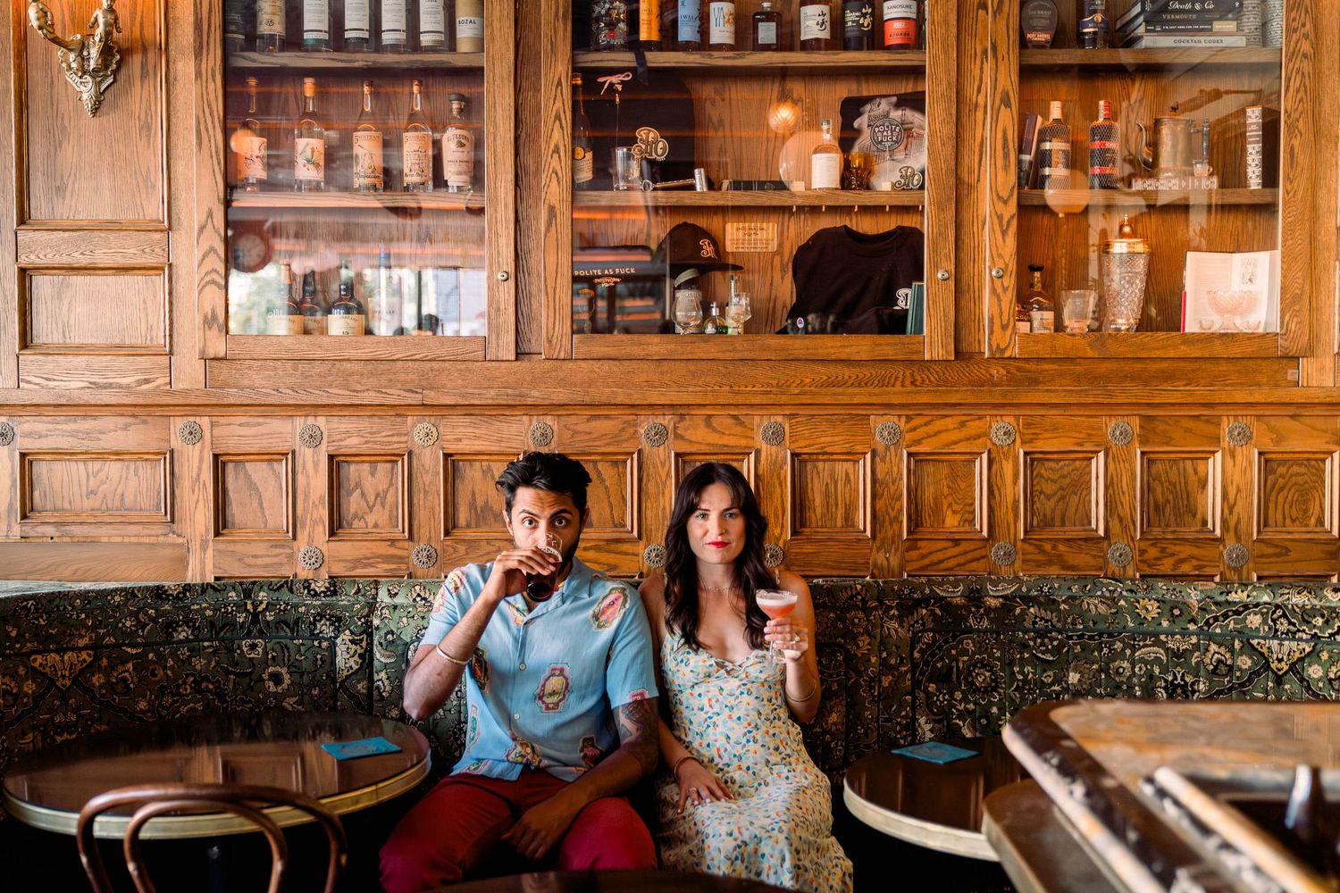 Couple sitting in a vintage restaurant with wooden cabinets and shelving displaying bottles and glassware in the background.