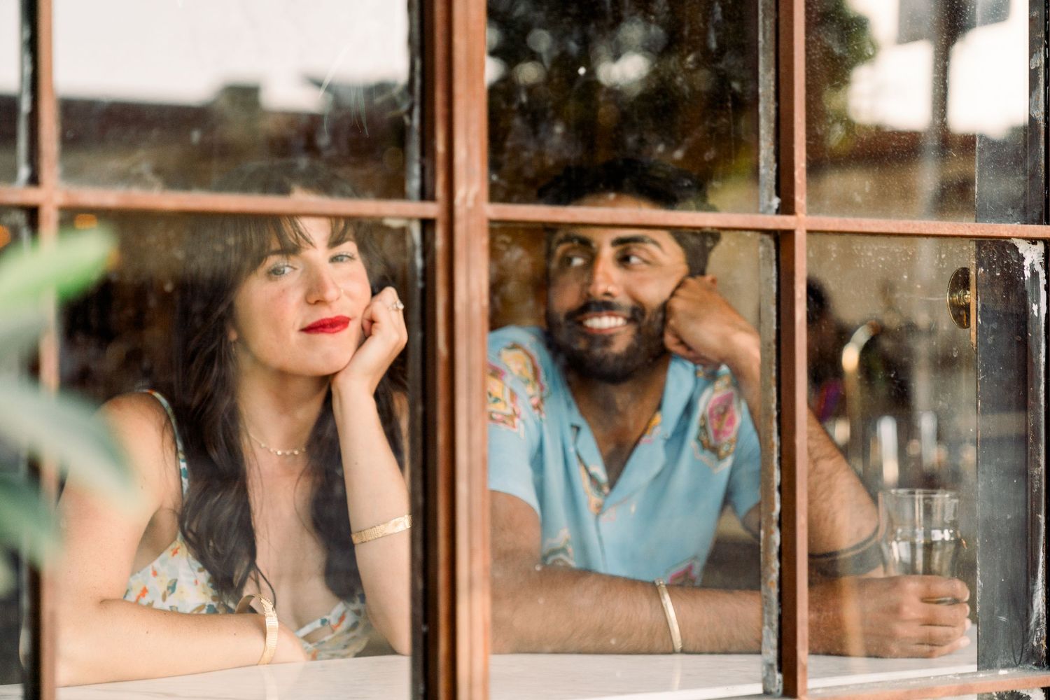Two people smile and pose playfully while looking through a window pane on a sunny day.