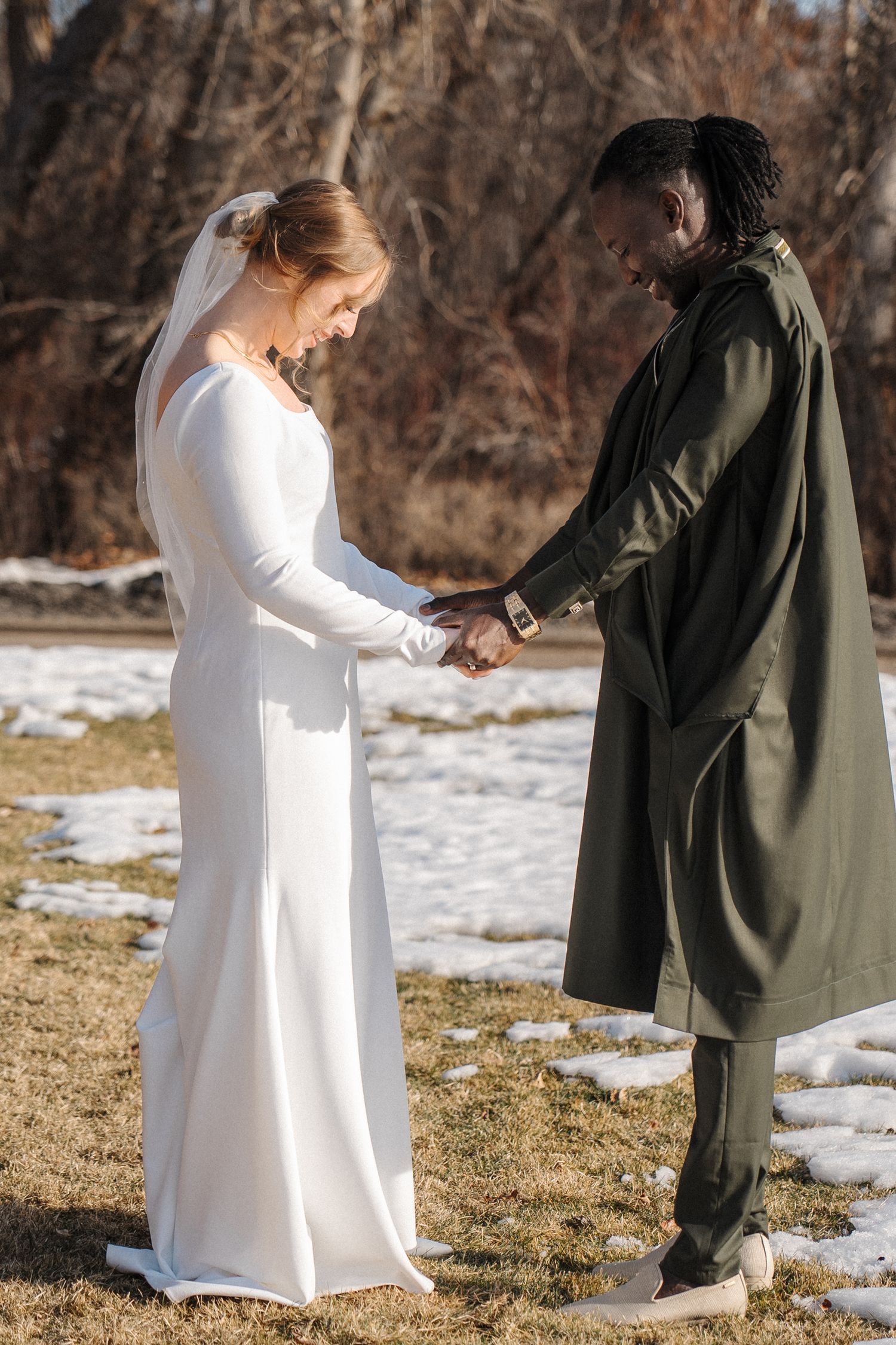A couple holds hands during their winter wedding ceremony with patches of snow on the ground in a natural outdoor setting.