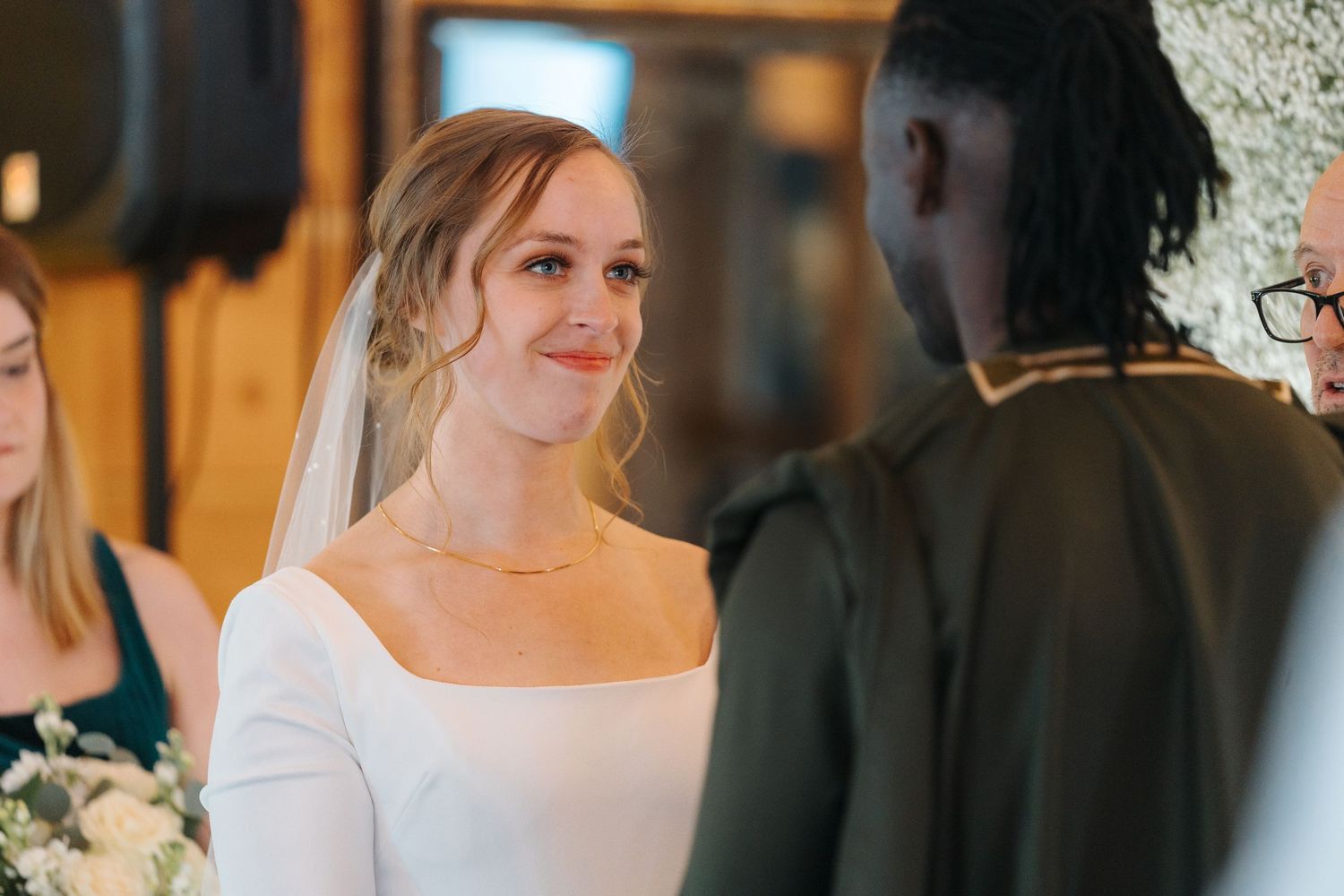 A bride in a white square-neck wedding dress exchanges vows during an intimate wedding ceremony indoors.