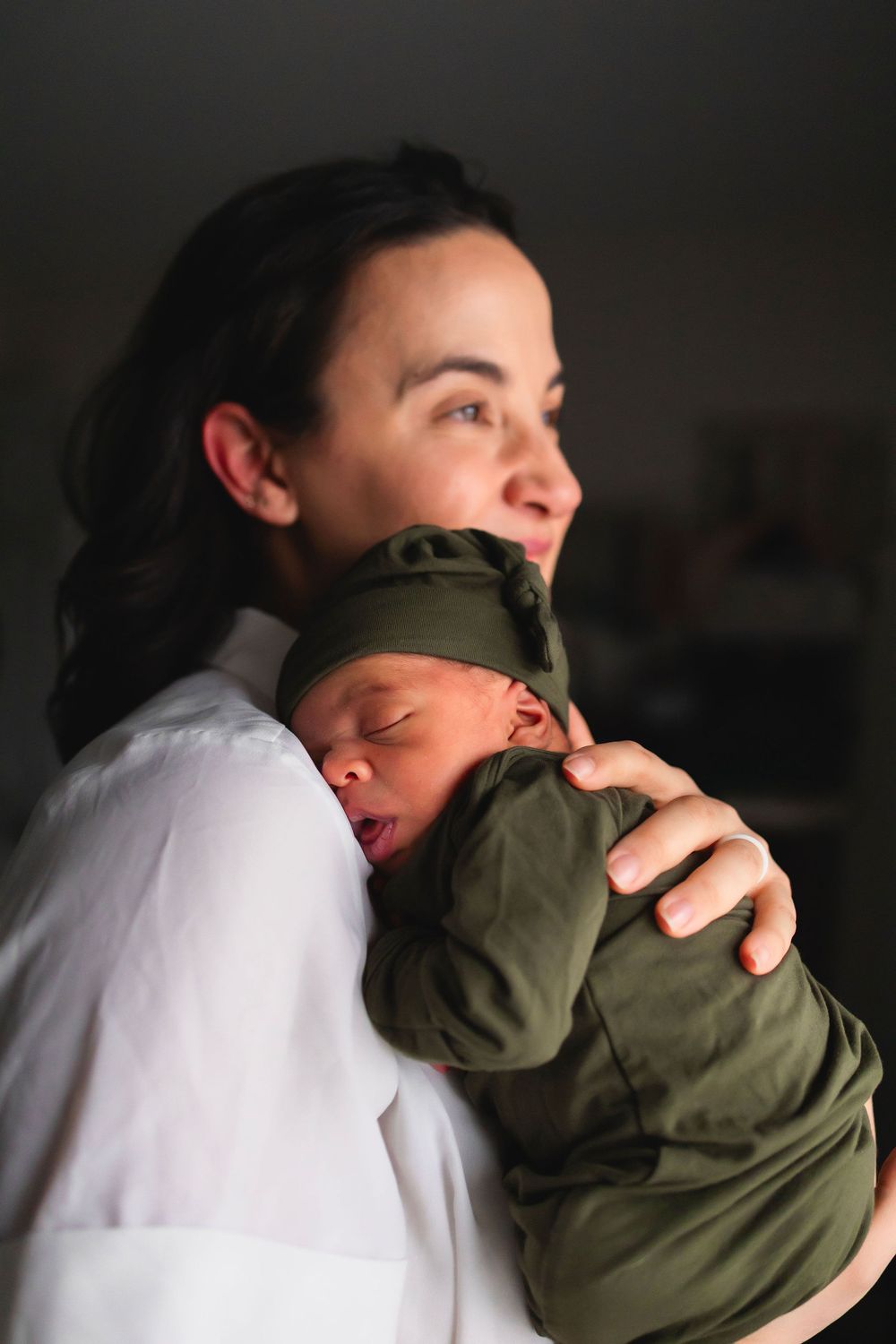 A sleeping baby in an olive green outfit rests peacefully against someone wearing a white shirt in a tender moment.